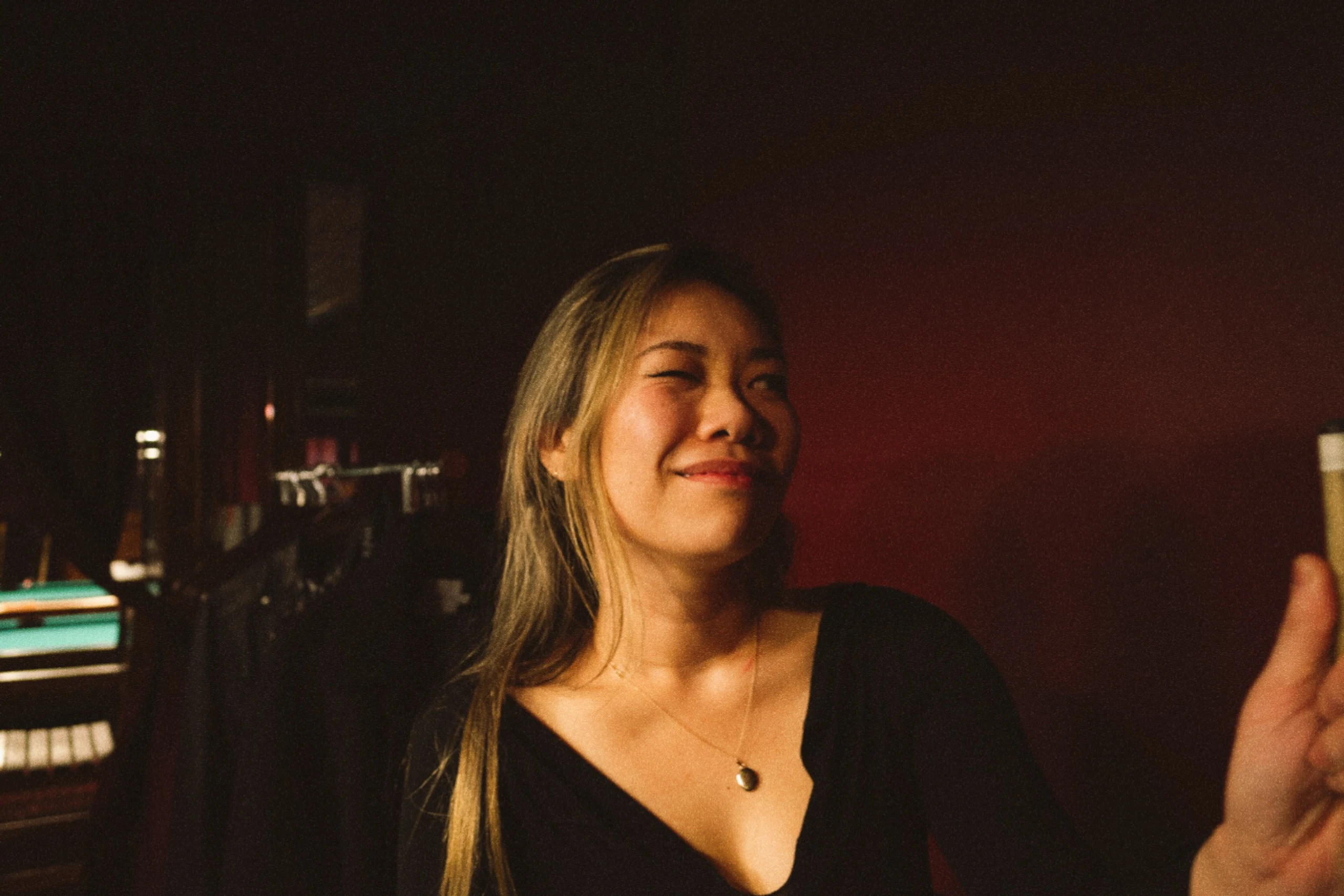 A woman smiles with her eyes closed in warm red pool-hall light.
