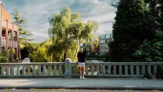A man stands centered against a stone canal bridge with water, trees, and brick buildings behind him in Amsterdam.