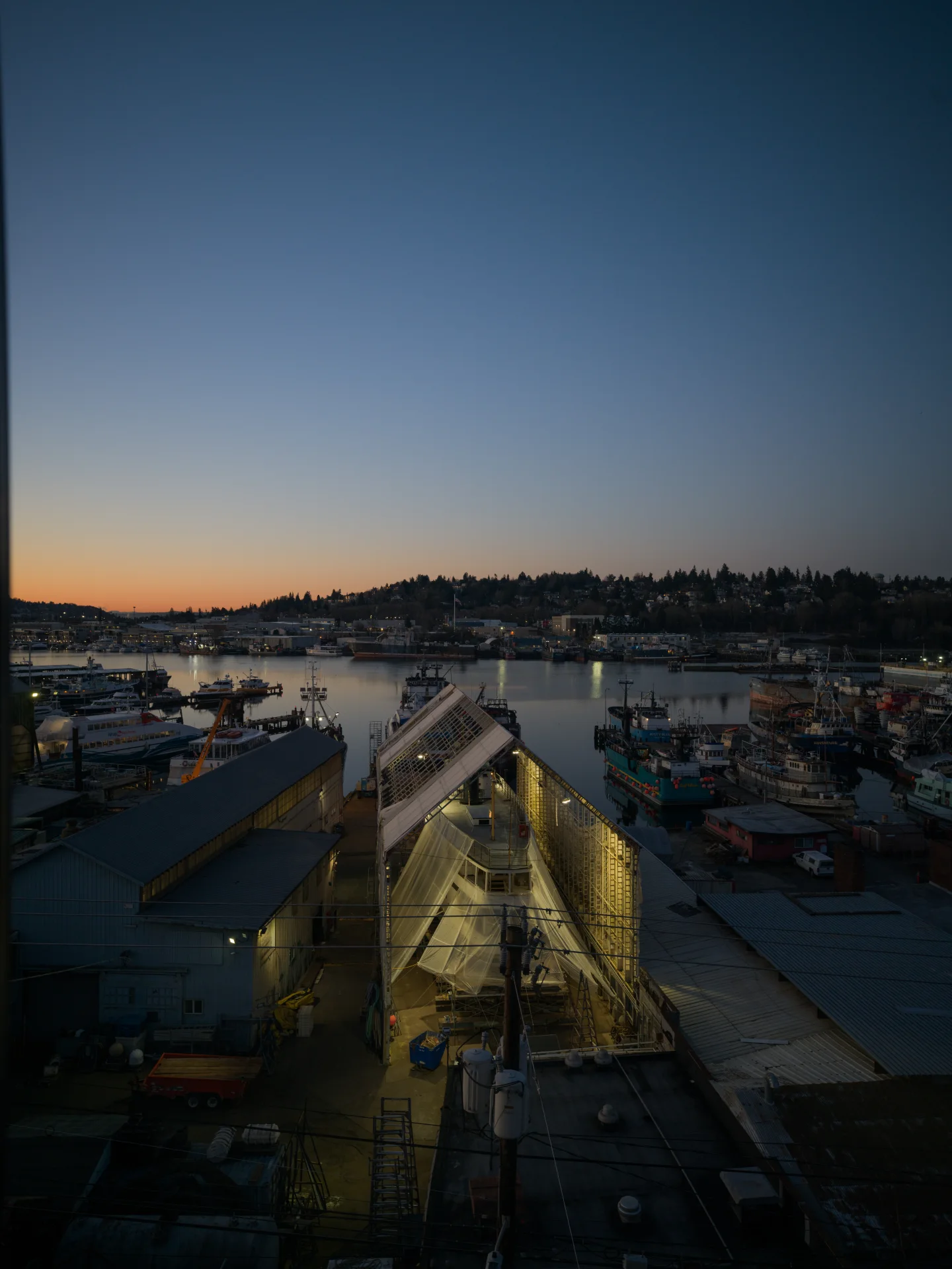 Boats and shipyard buildings fill a Seattle harbor at dusk, with work lights glowing beneath a blue-and-orange sky.