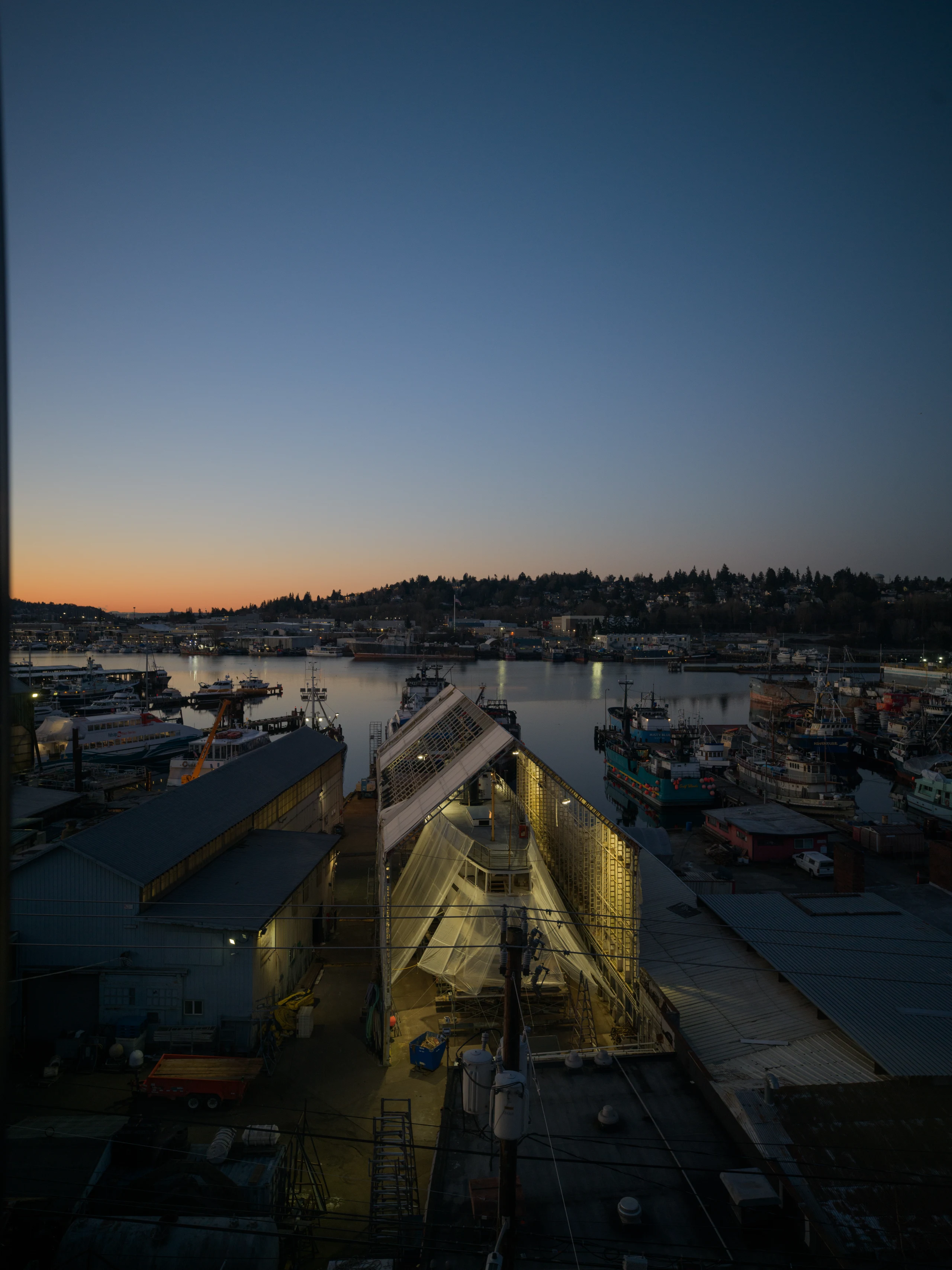 Boats and shipyard buildings fill a Seattle harbor at dusk, with work lights glowing beneath a blue-and-orange sky.