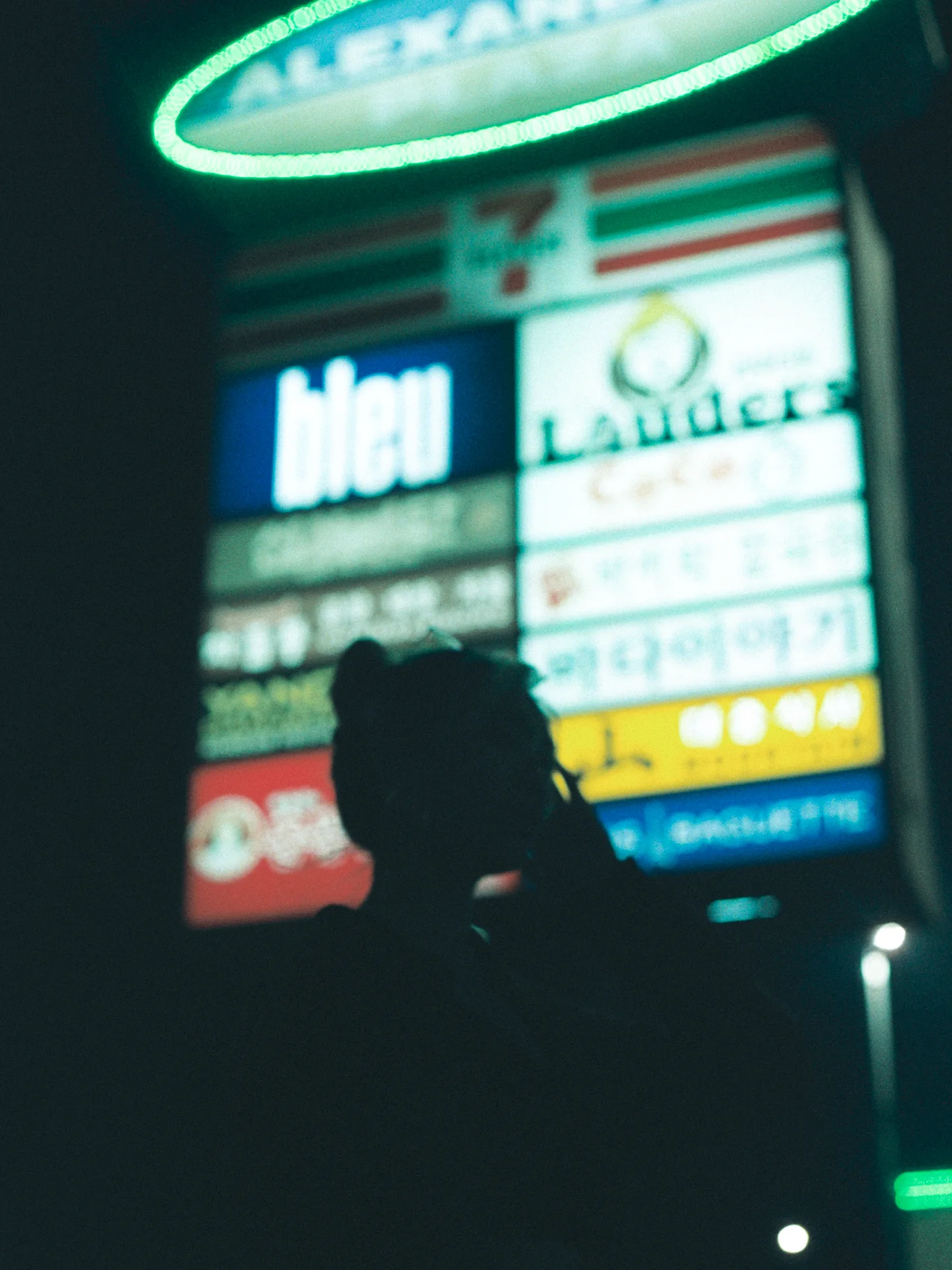 A blurred silhouetted person passes beneath green and blue illuminated storefront signs at night.