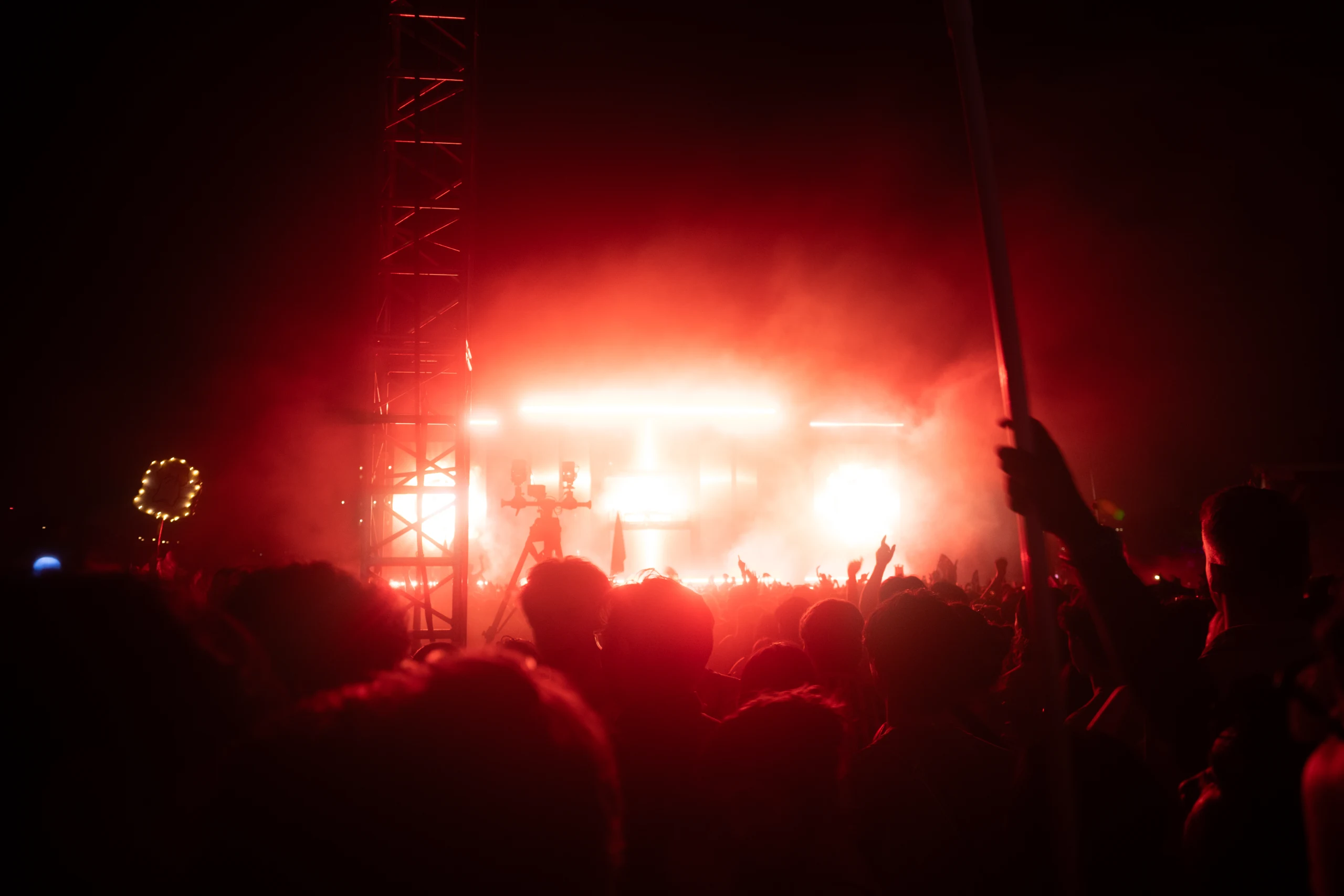 Concert crowd silhouetted against a bright red stage with smoke, raised hands, and a production tower.