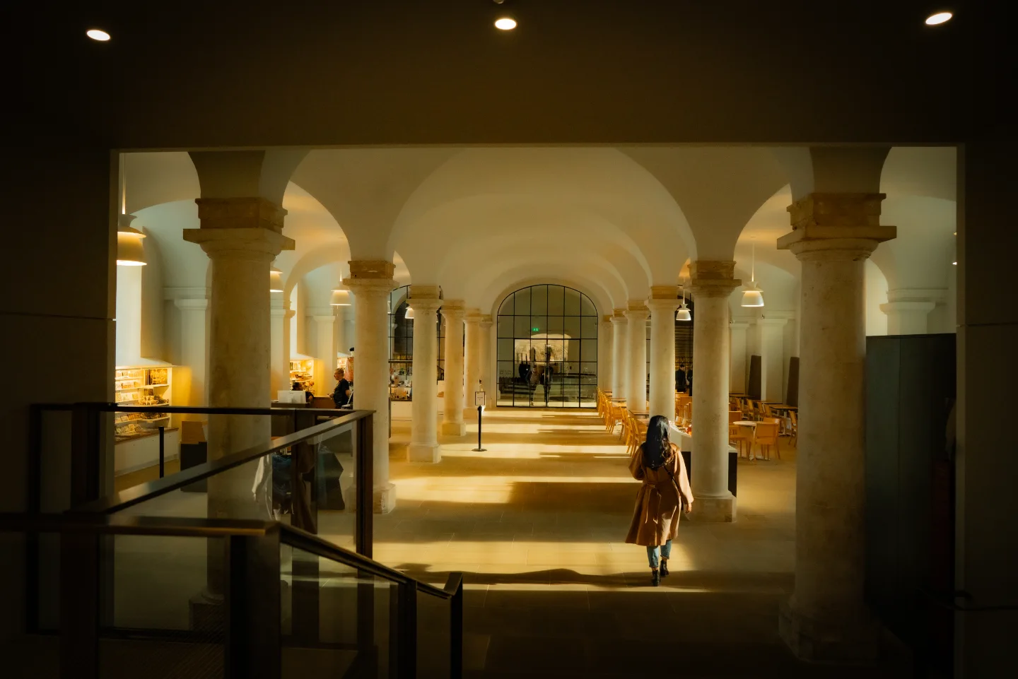A lone visitor walks through a warm sunlit arcade of columns and arches.