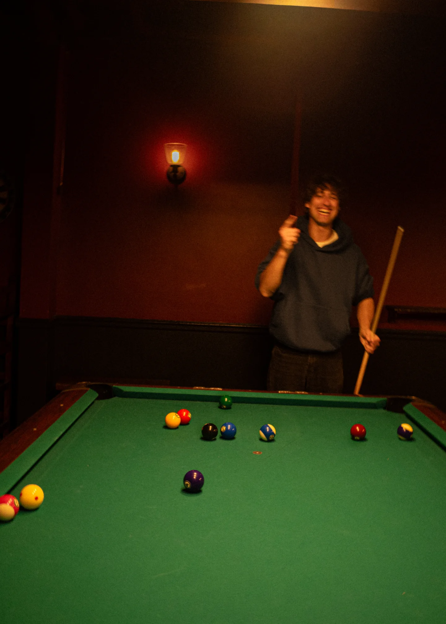 A pool player holds a cue beneath a square overhead lamp against a red wall.