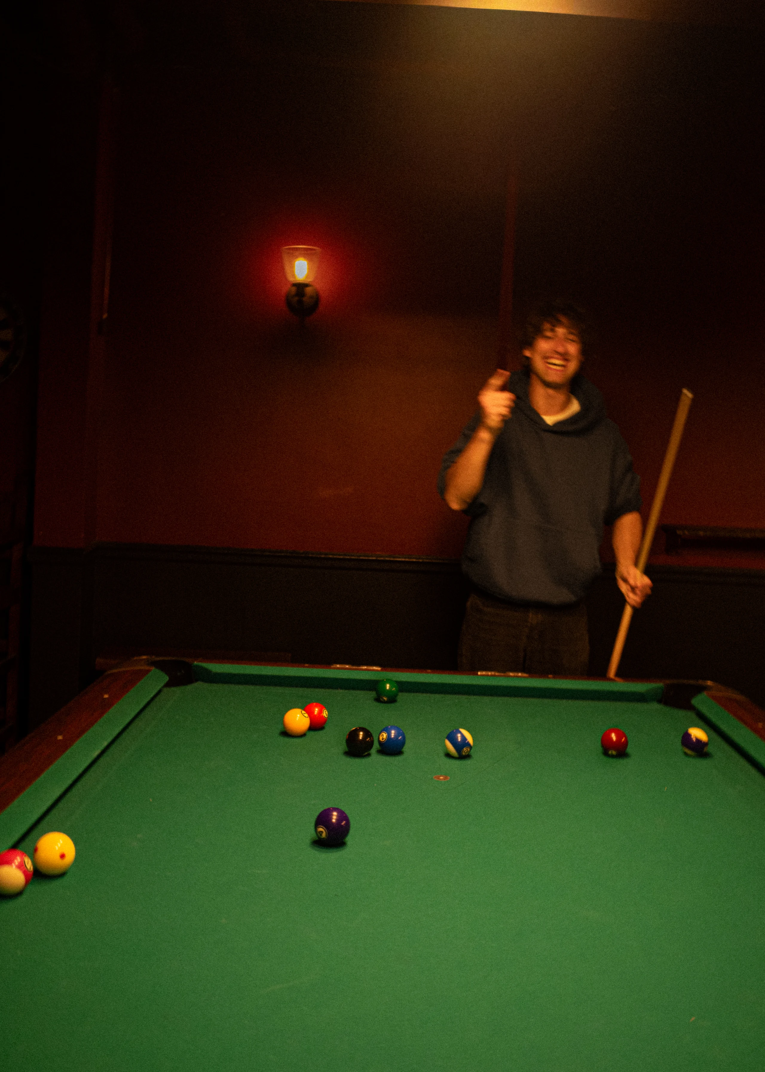 A pool player holds a cue beneath a square overhead lamp against a red wall.
