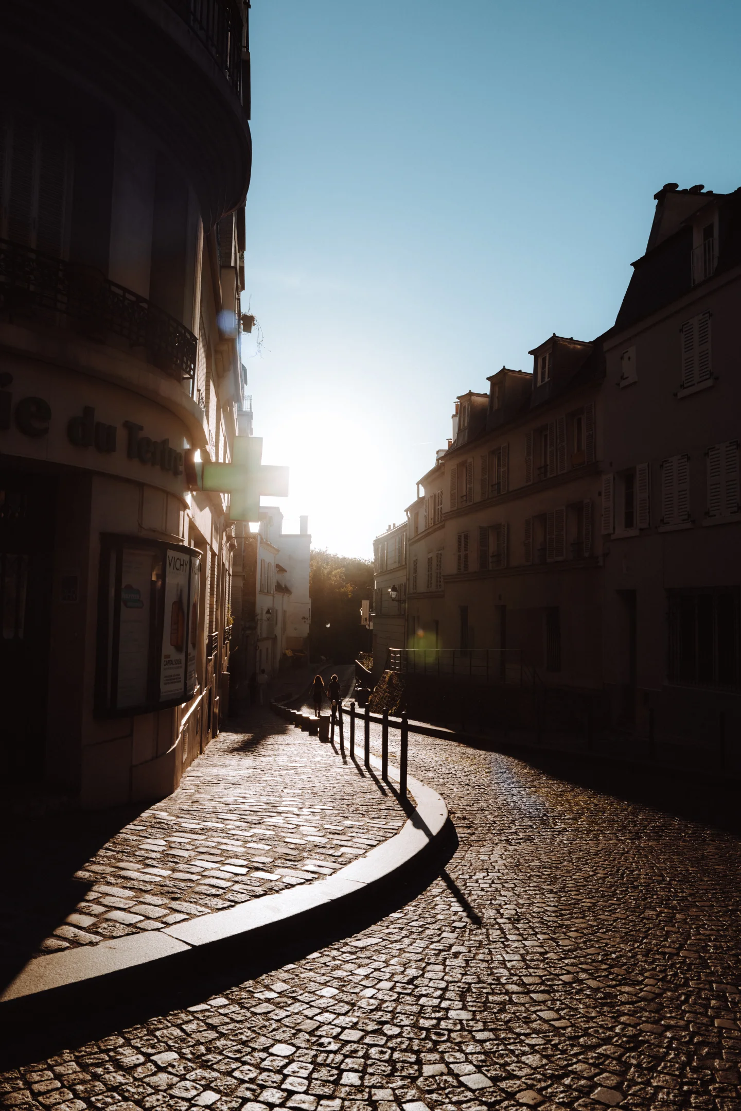 Low afternoon sun silhouettes two people walking down a curved cobblestone street between dark buildings in Montmartre.