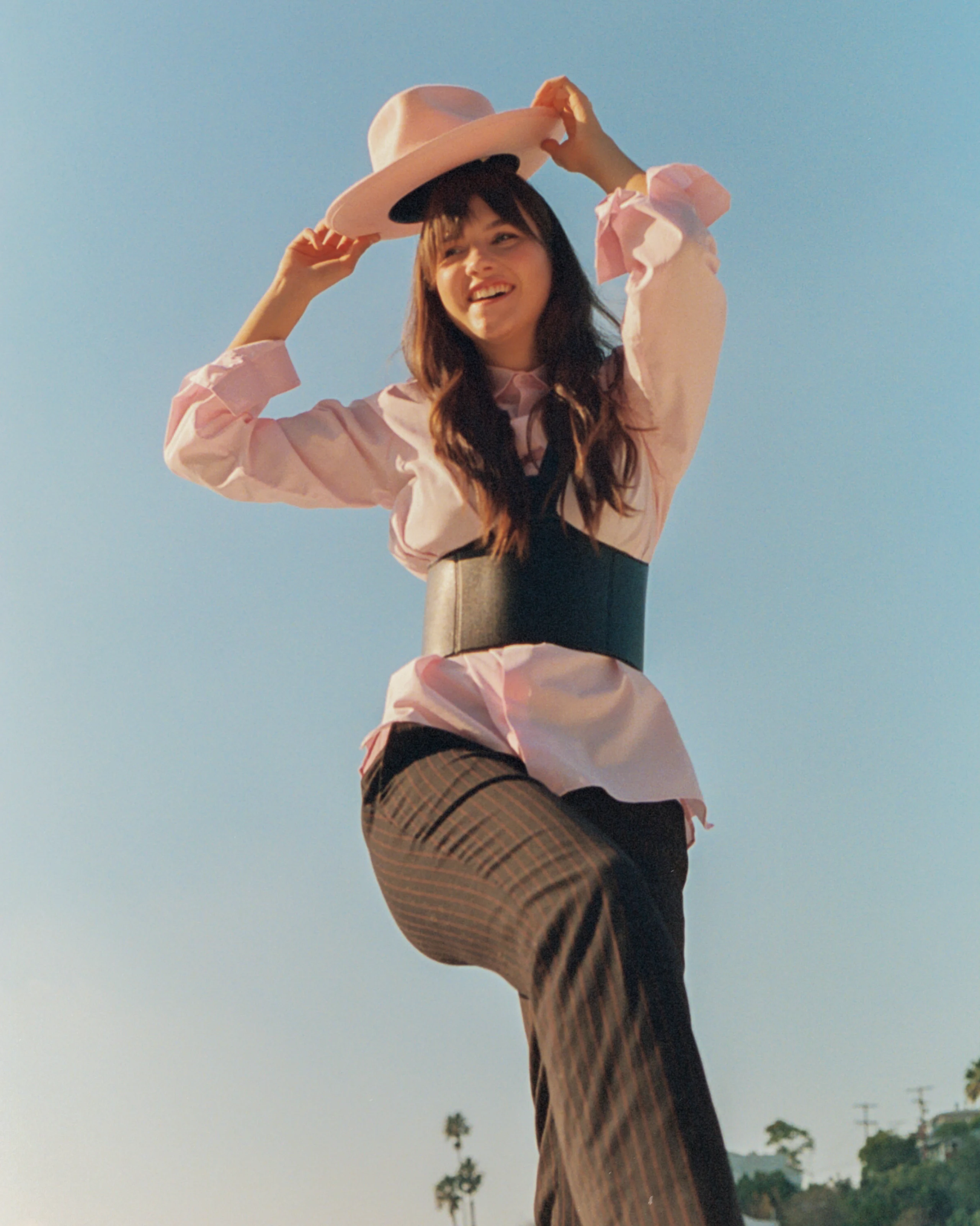 A woman in a pale pink hat and layered clothing posing against a clear blue sky.