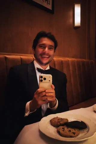 A smiling man in a tuxedo holding a compact camera at a dinner table.