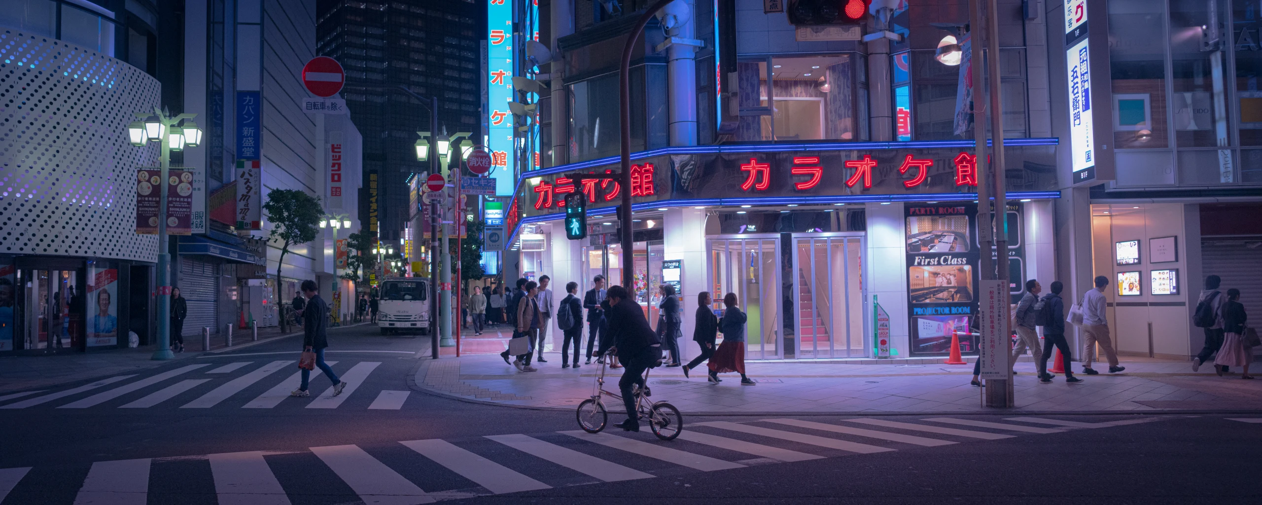 Pedestrians cross a blue-lit entertainment street at night.