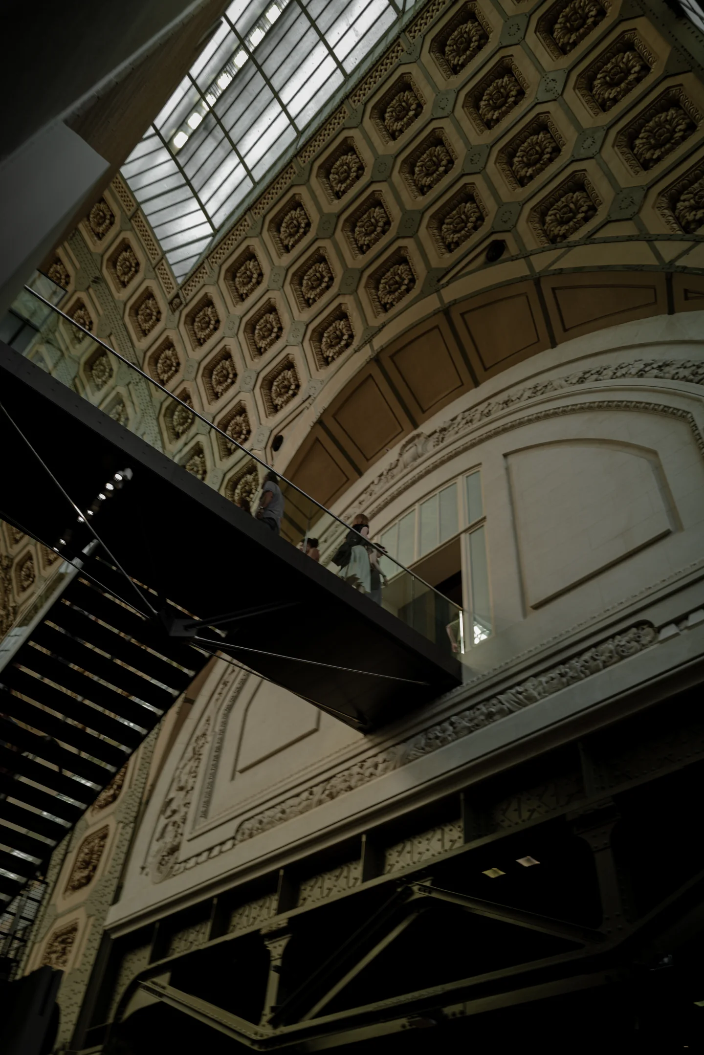 Black stairways crossing an ornate museum wall and glass roof, viewed from below.