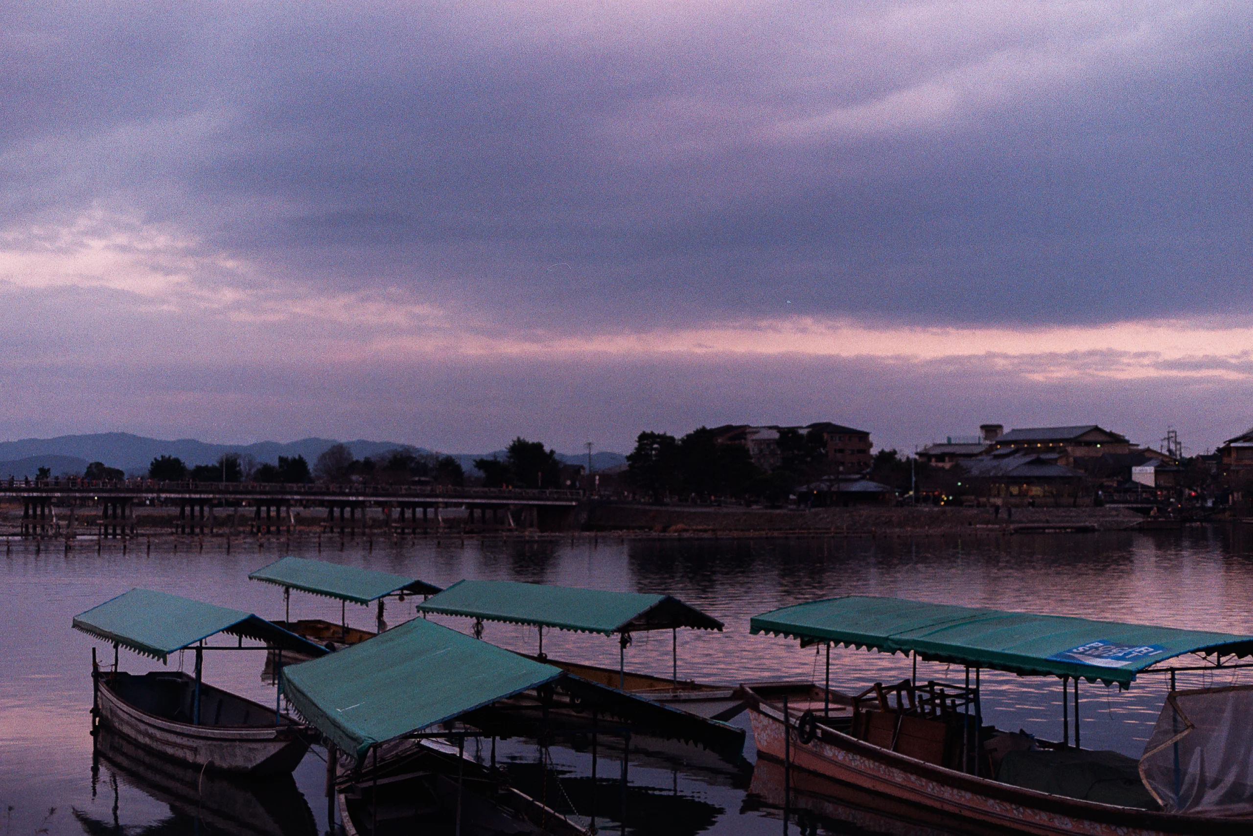 Covered boats rest beside a calm river under a violet sky.