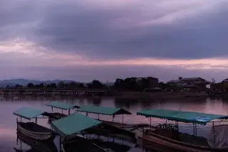 Covered boats rest beside a calm river under a violet sky.