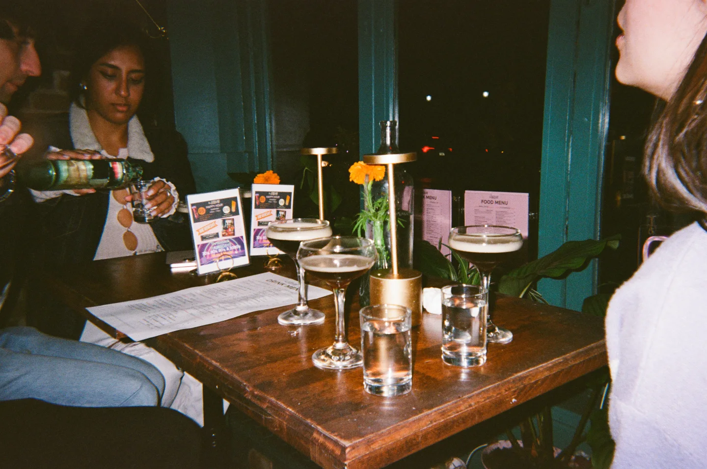 Friends sit around a restaurant table crowded with glasses and small lamps.