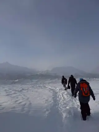 Three bundled hikers walk single file across a windswept snowfield toward hazy Alaska mountains.