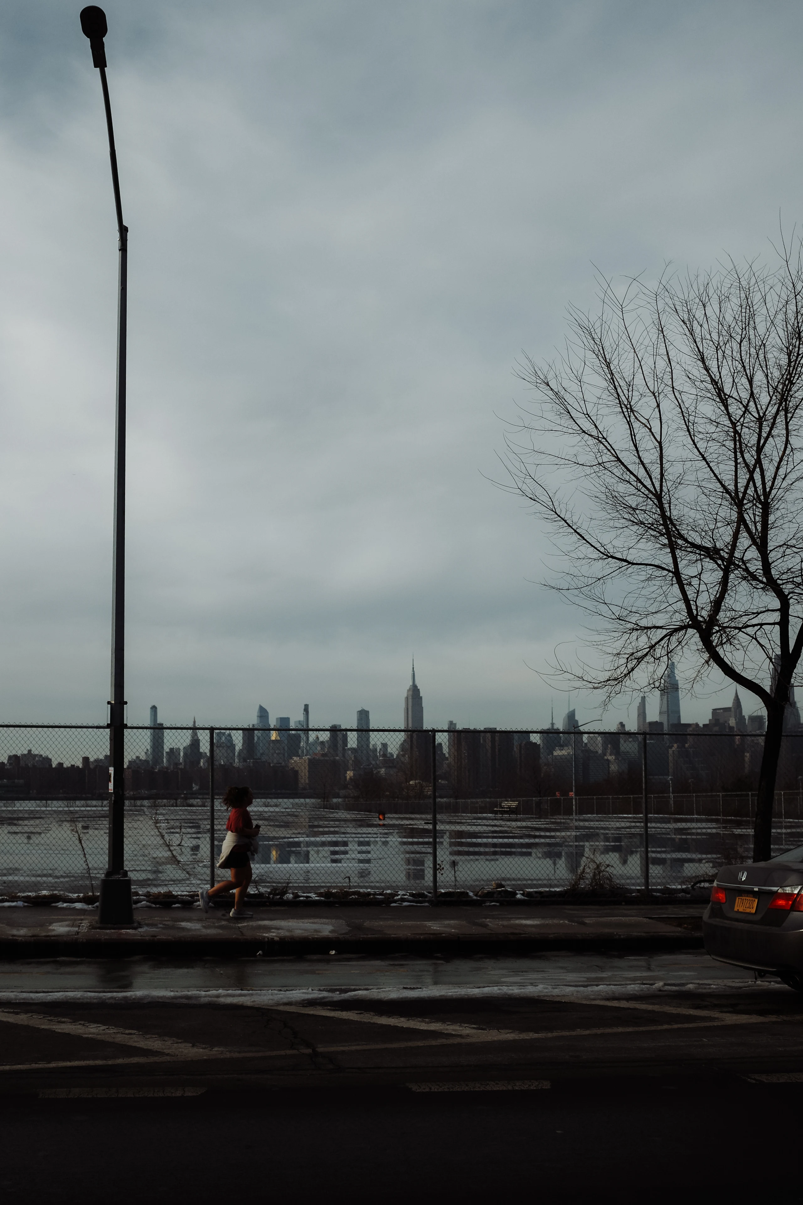 A runner passes a fenced waterfront beneath a gray sky, with the Manhattan skyline across the river.