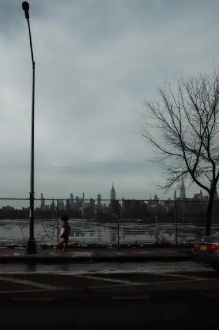 A runner passes a fenced waterfront beneath a gray sky, with the Manhattan skyline across the river.