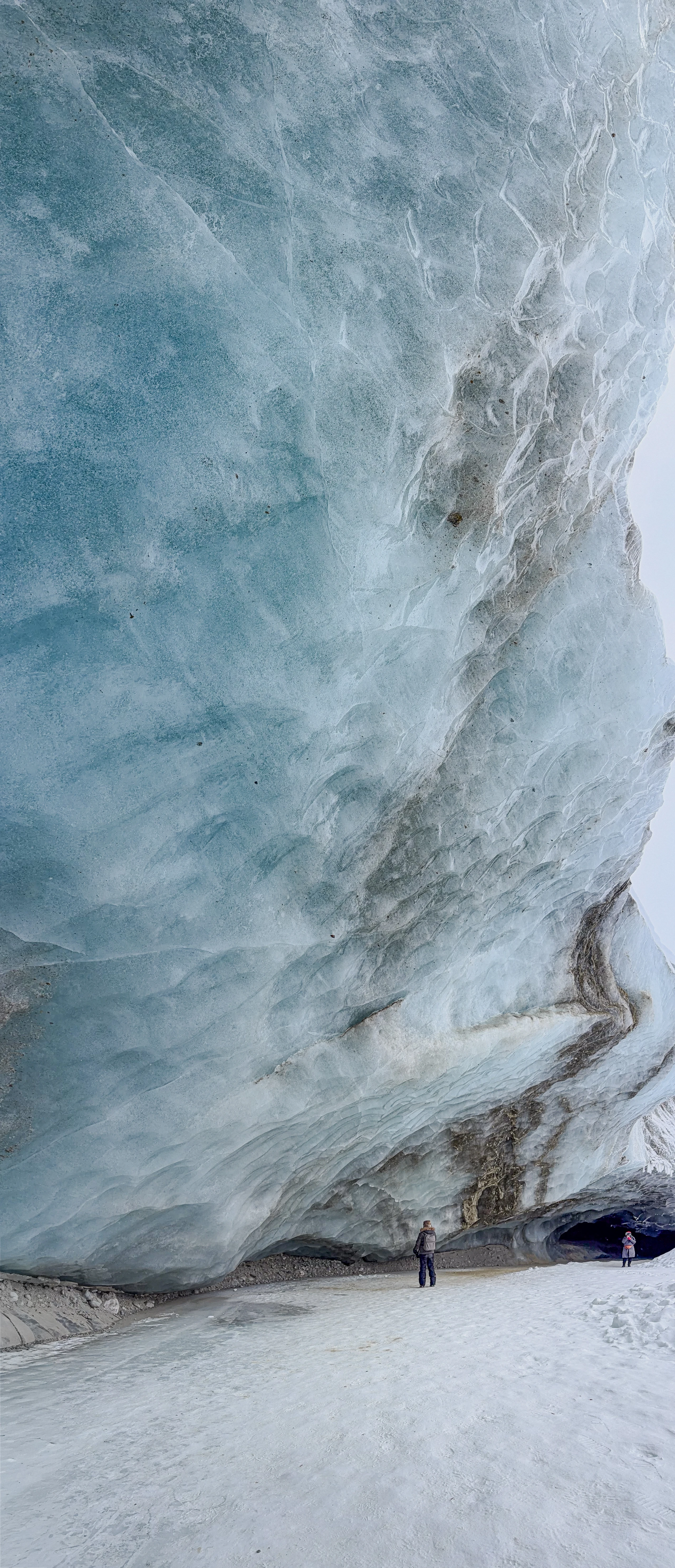 Two visitors stand beneath a towering blue glacier wall streaked with rock and layered ice.
