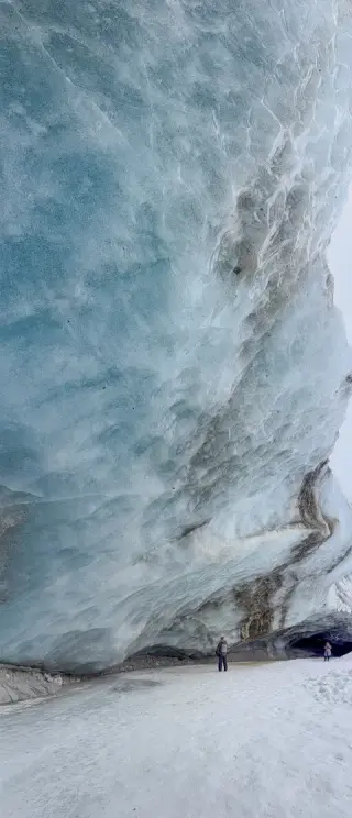 Two visitors stand beneath a towering blue glacier wall streaked with rock and layered ice.