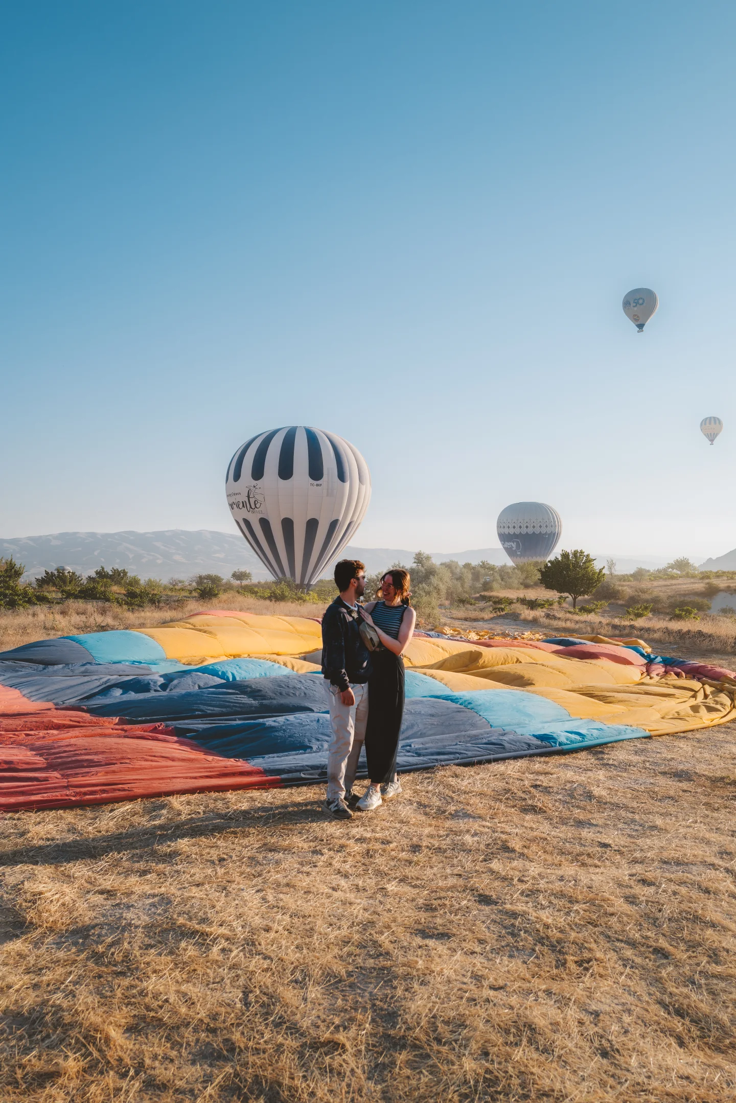 Two people stand beside colorful deflated balloon fabric as hot-air balloons rise beyond them.