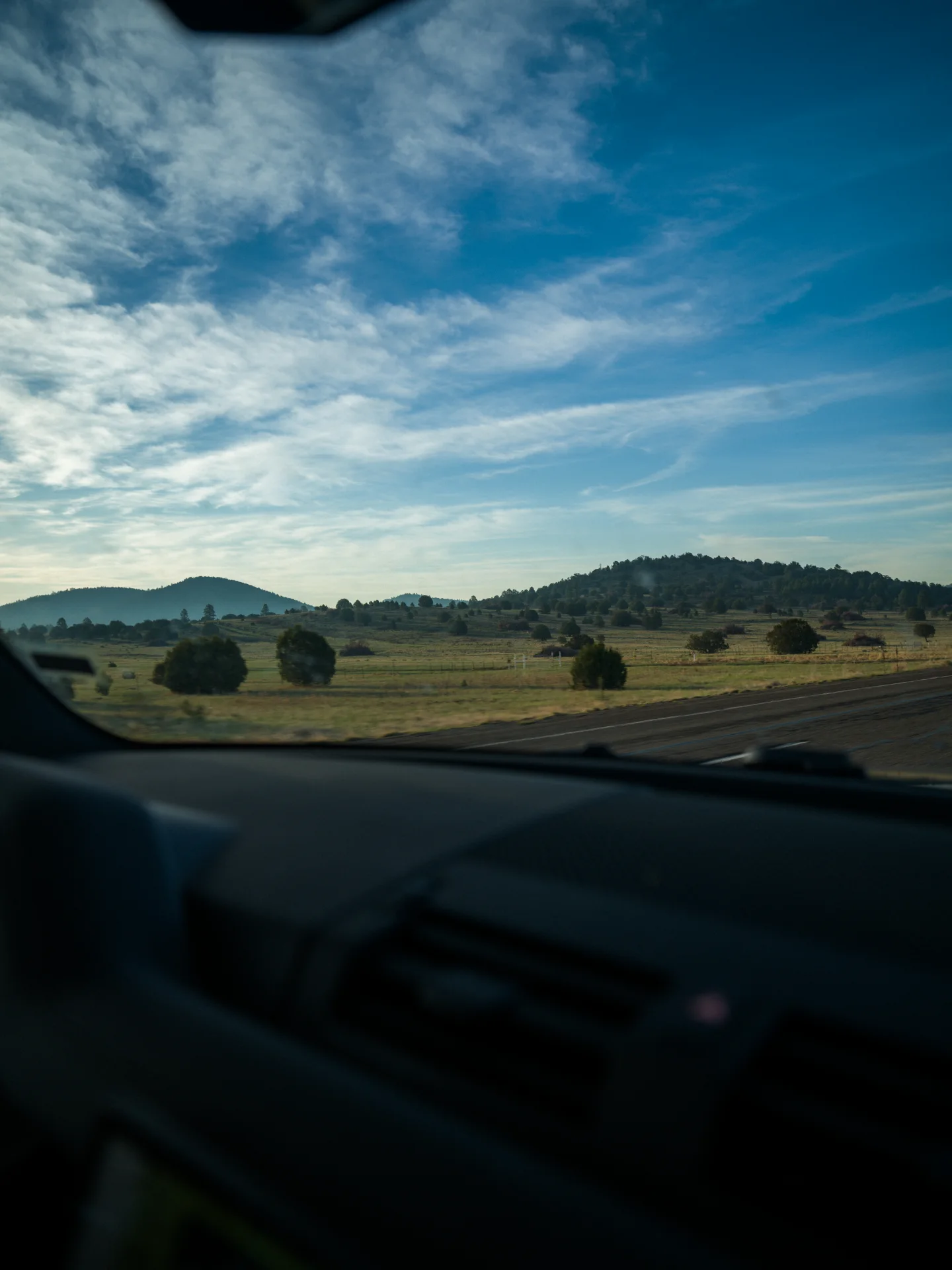 Juniper-dotted grassland and low hills seen through a car windshield beneath a blue sky.