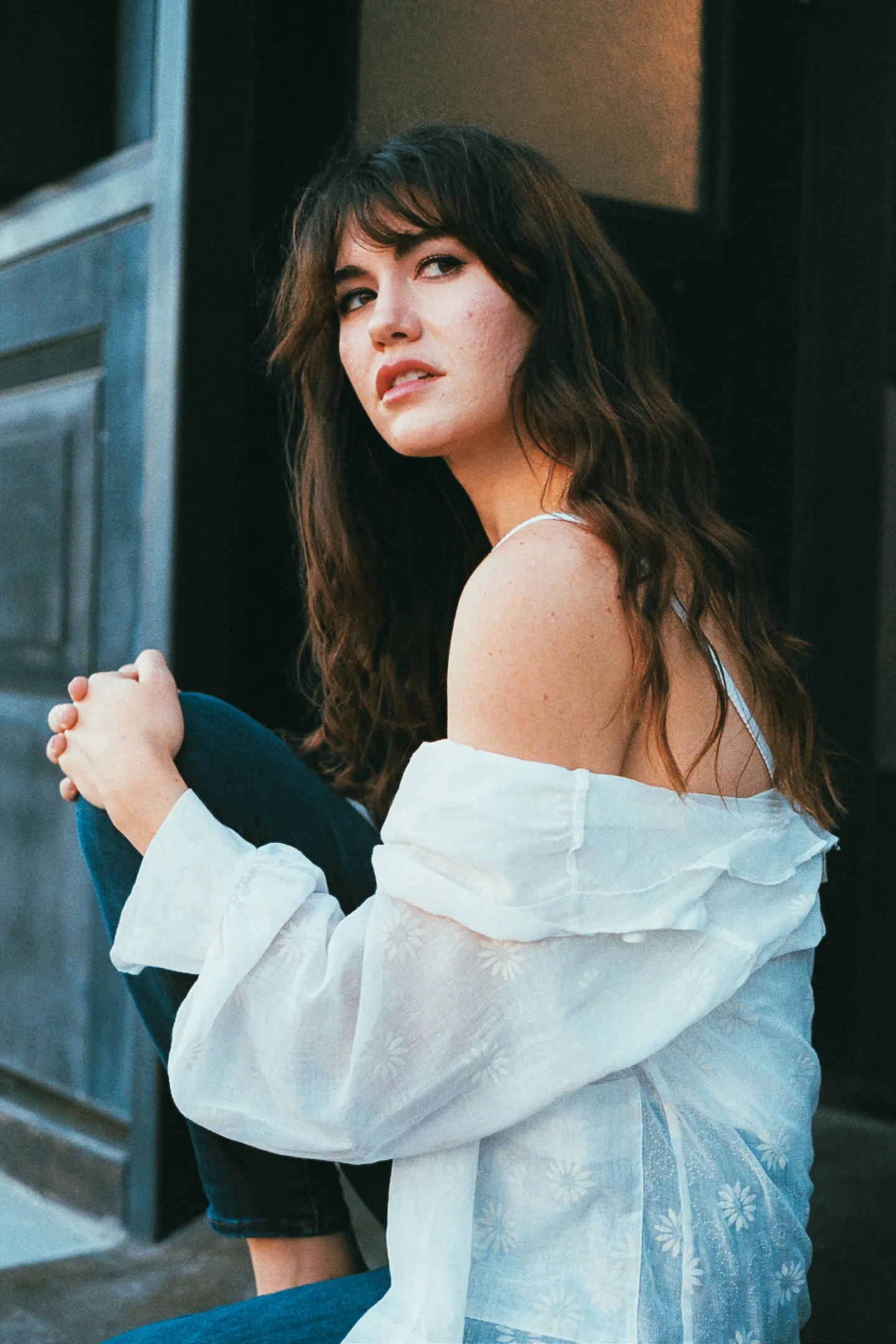 A seated woman with long dark hair looks upward while wearing a white blouse beside a blue-gray doorway.