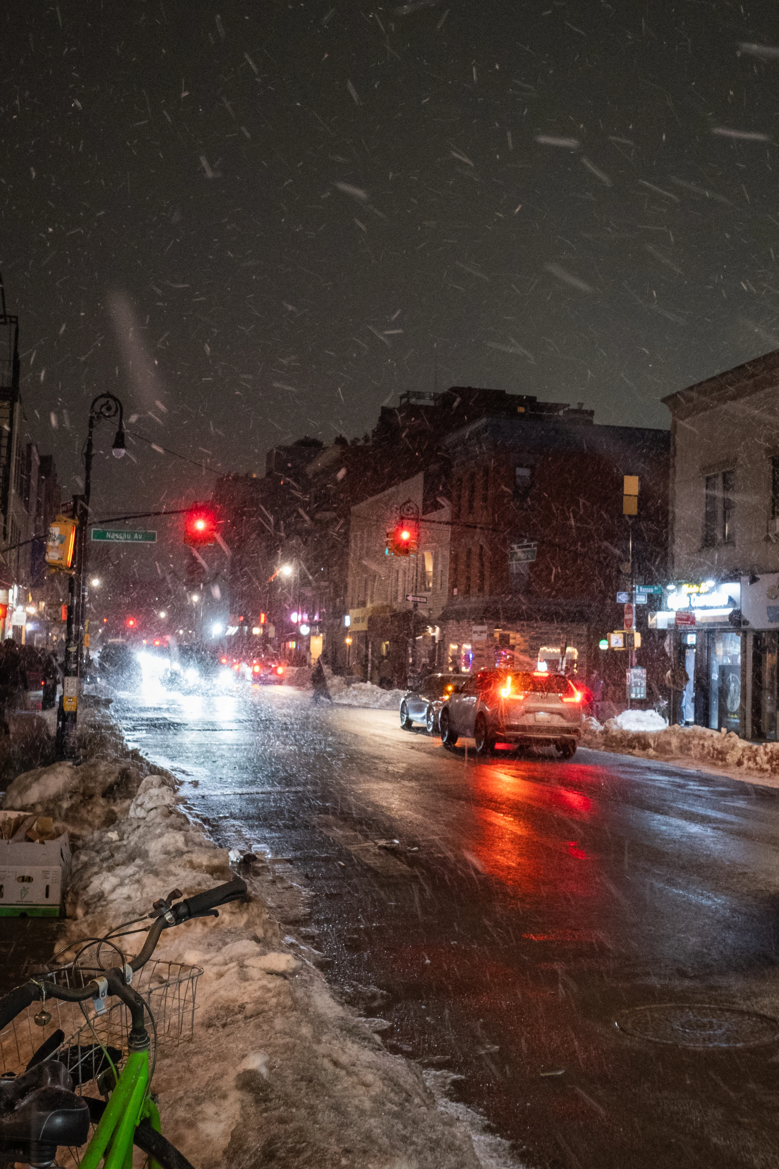 A wet snowy city street at night with cars and red traffic lights.