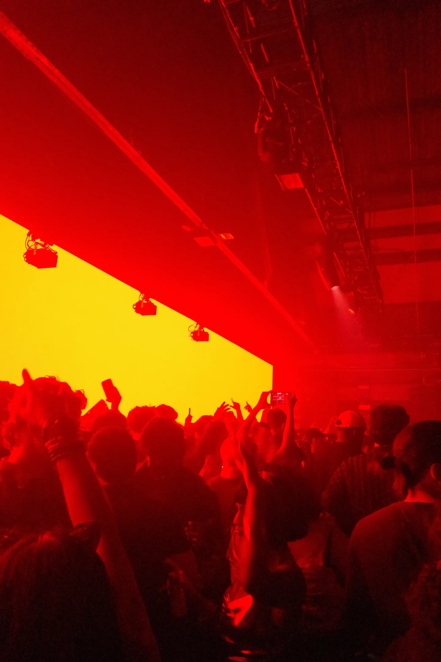 Crowd bathed in red light facing a large yellow screen inside a music venue.