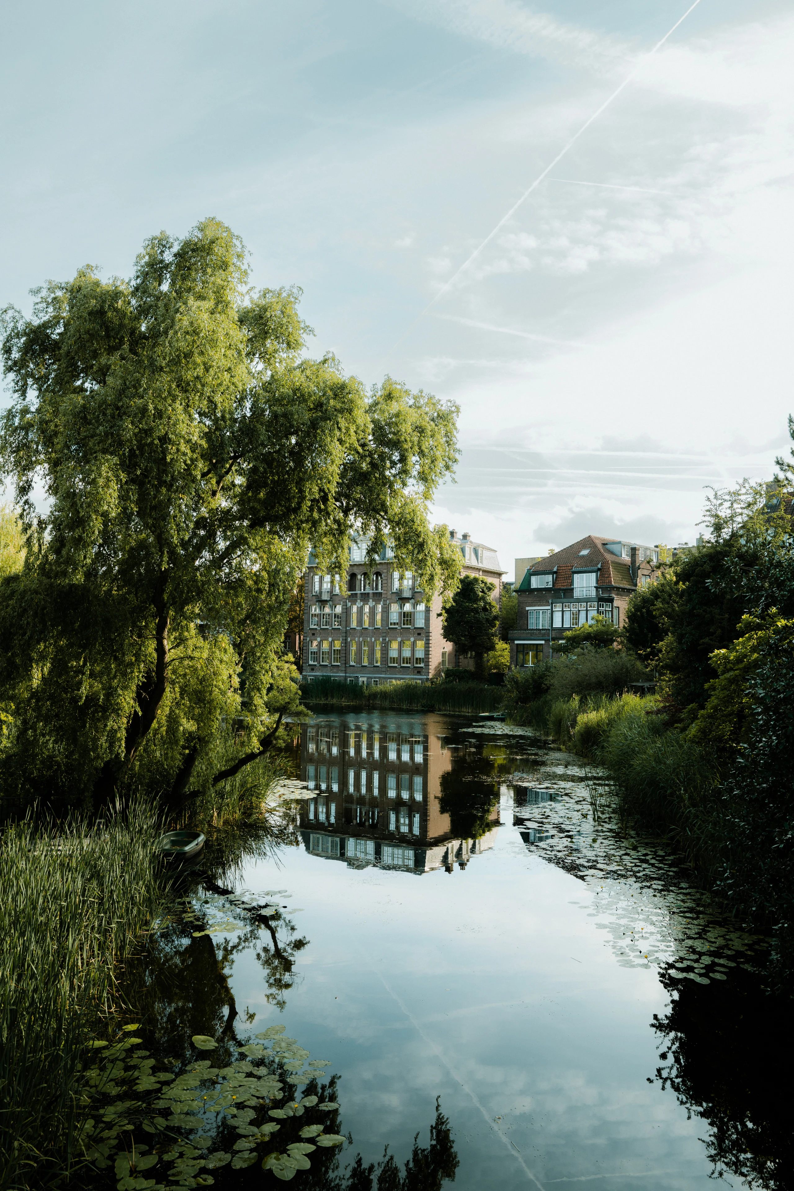 Willow trees and residential buildings reflected in a narrow Amsterdam pond under a blue sky.