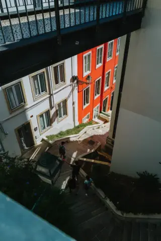 People descend a stairway beneath a dark footbridge, between white and orange building façades.
