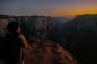 A person stands at a Zion canyon overlook as warm sunset light fades.