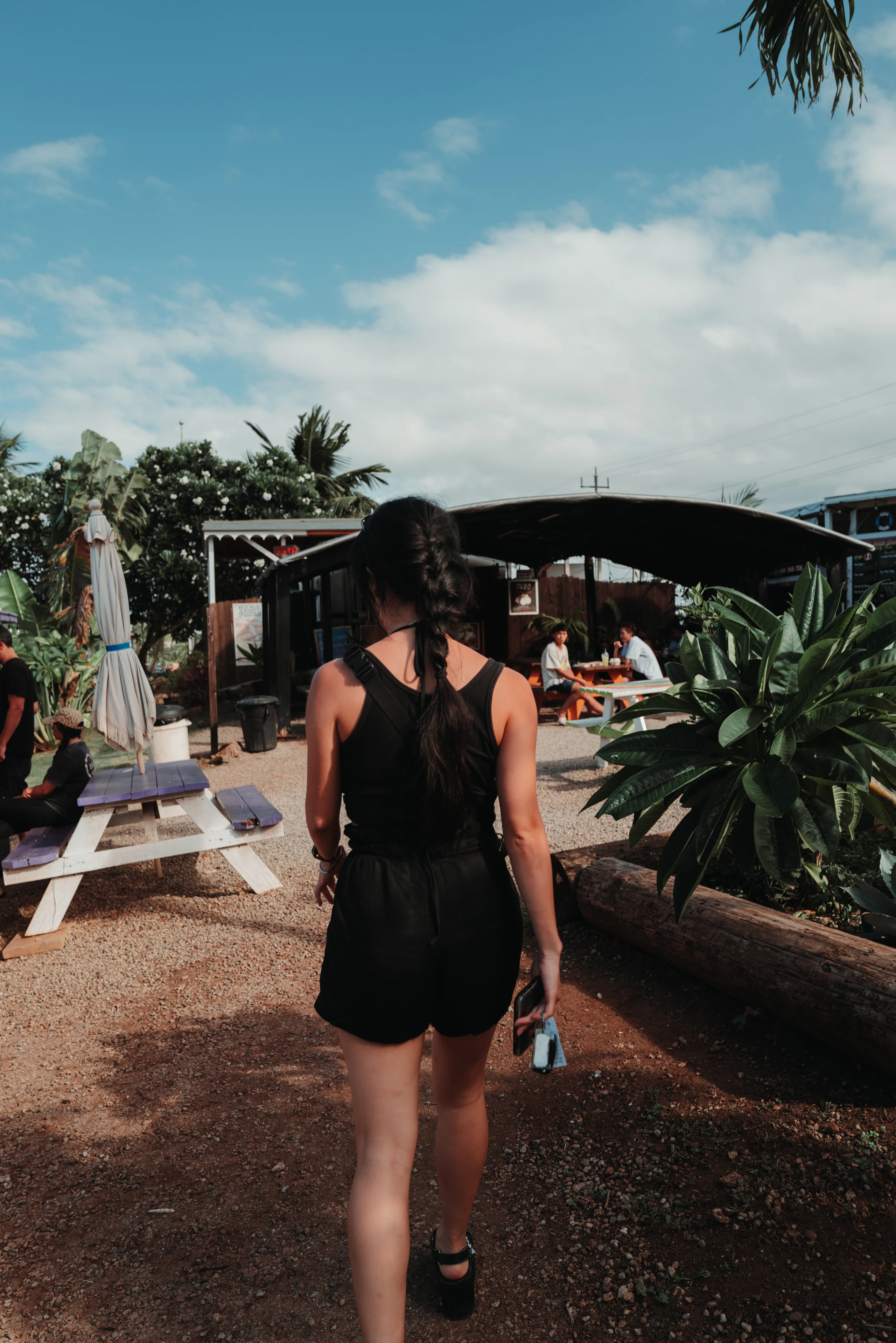 A woman with a long braid walks between picnic tables and tropical plants beneath a blue sky.