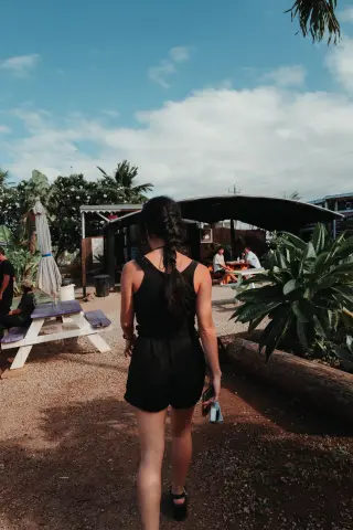A woman with a long braid walks between picnic tables and tropical plants beneath a blue sky.