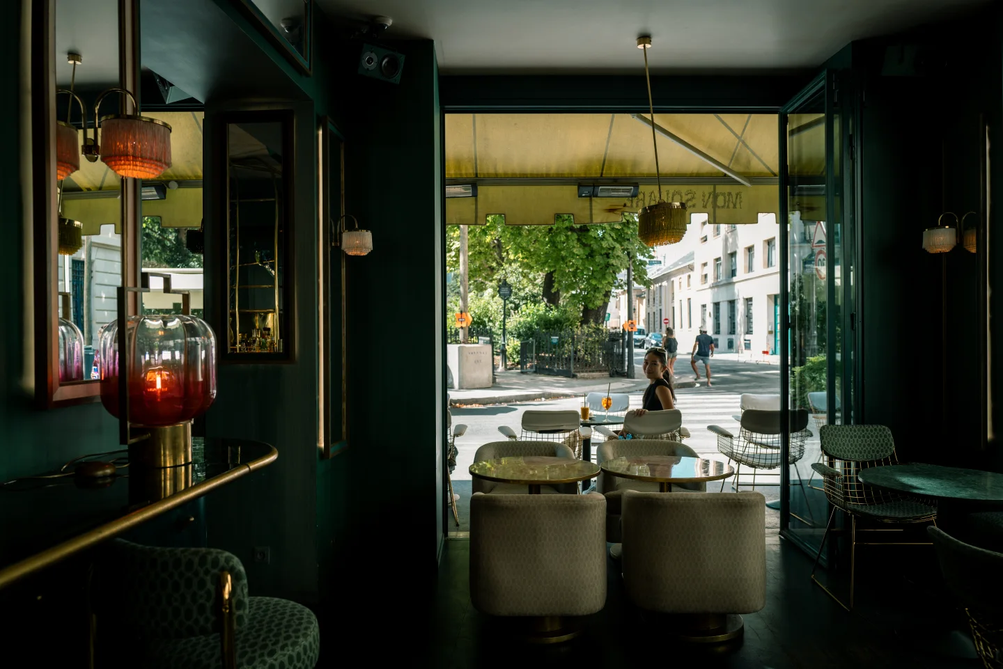 View from a dark Paris café through open doors to sidewalk tables, where a seated woman looks toward the camera.