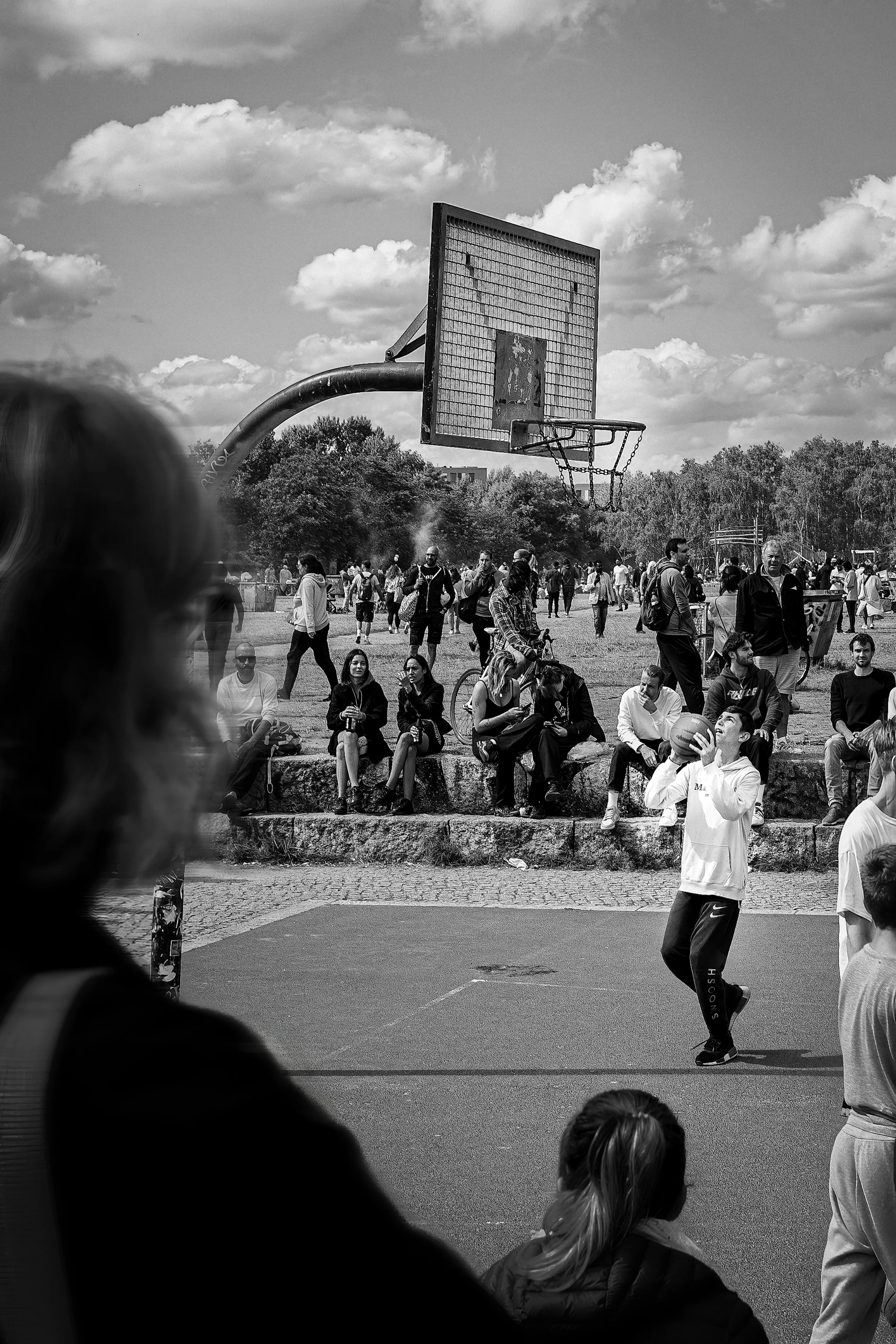 Black-and-white photograph of a basketball player beneath a weathered hoop as spectators gather at Mauerpark.