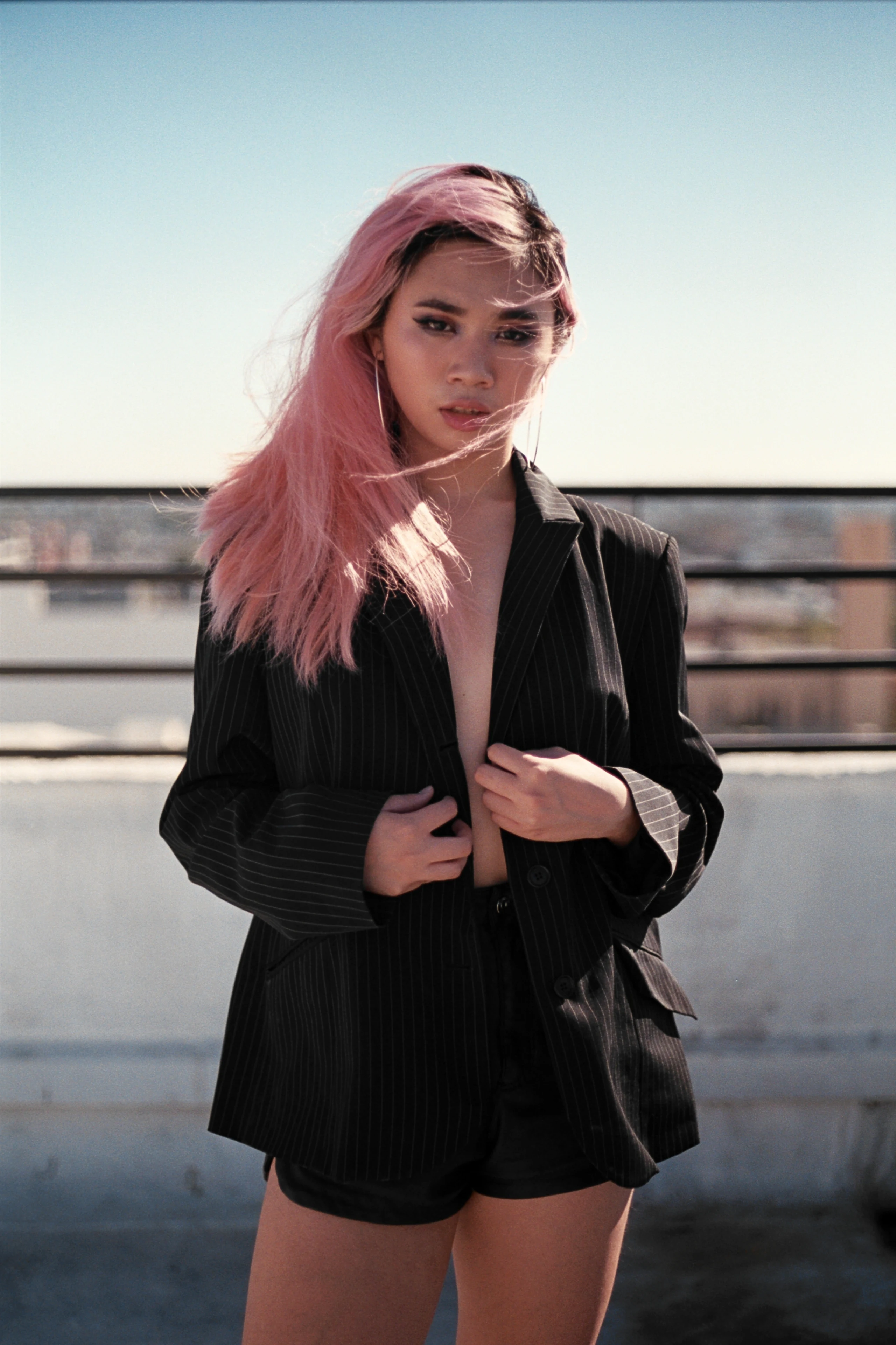A woman with pink hair wearing a black suit on a rooftop under a pale blue sky.