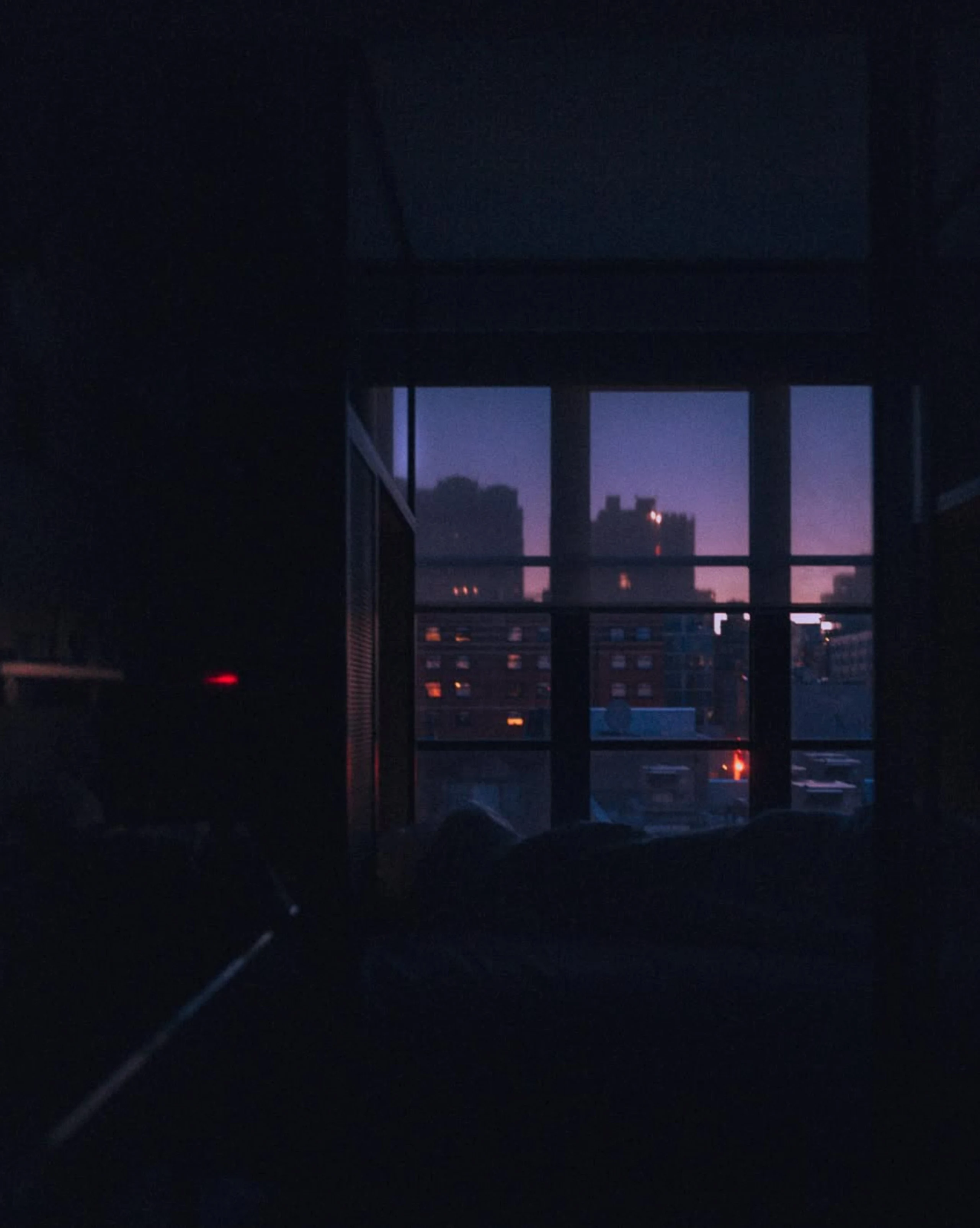 A dark interior frames a window looking onto New York apartment buildings beneath a violet dusk sky, with scattered lights glowing inside.