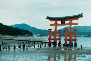 Visitors walk across an exposed tidal flat beneath a large red torii gate.