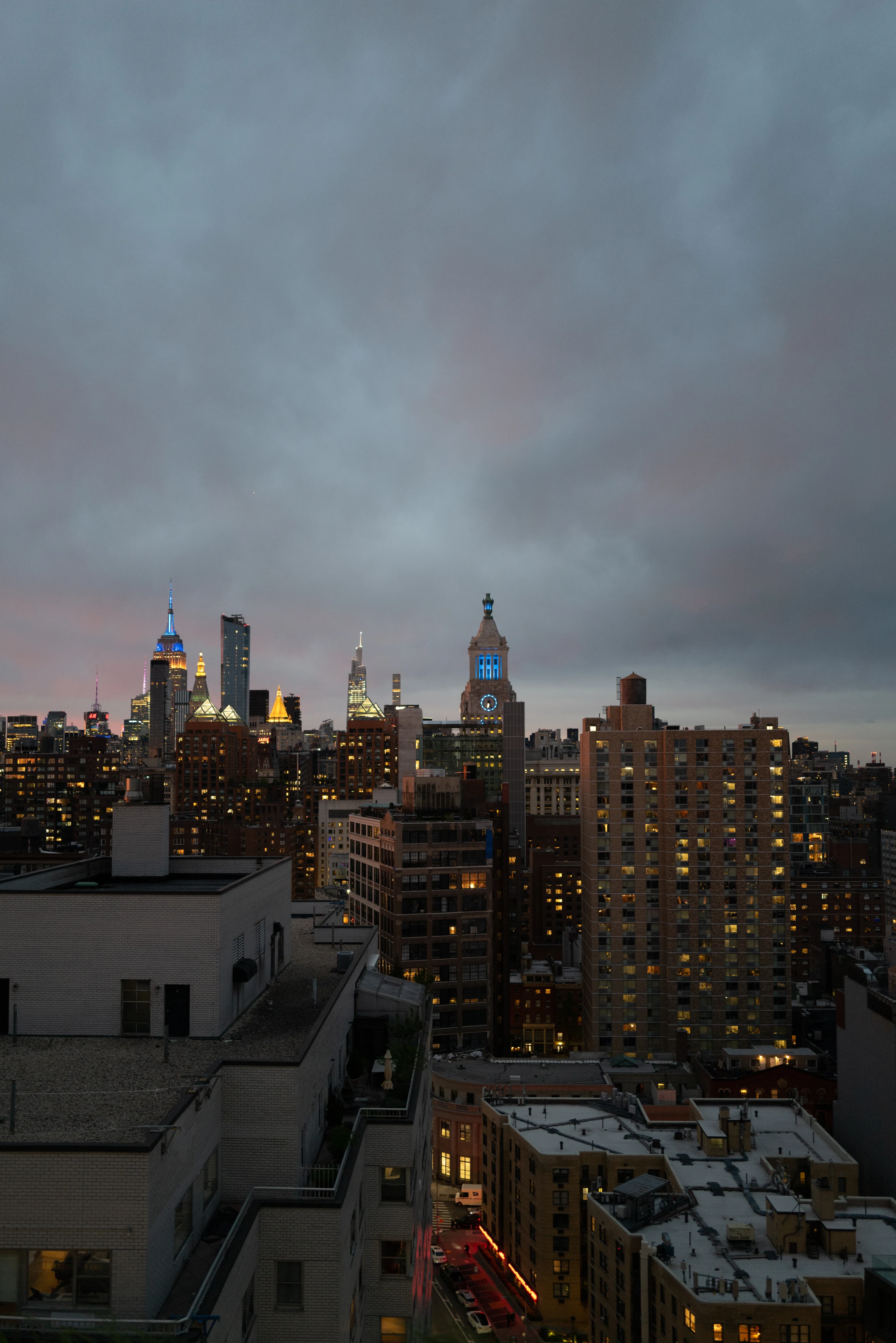 The Manhattan skyline at blue hour under heavy gray clouds, with the Empire State Building lit blue and gold above dense rooftops.