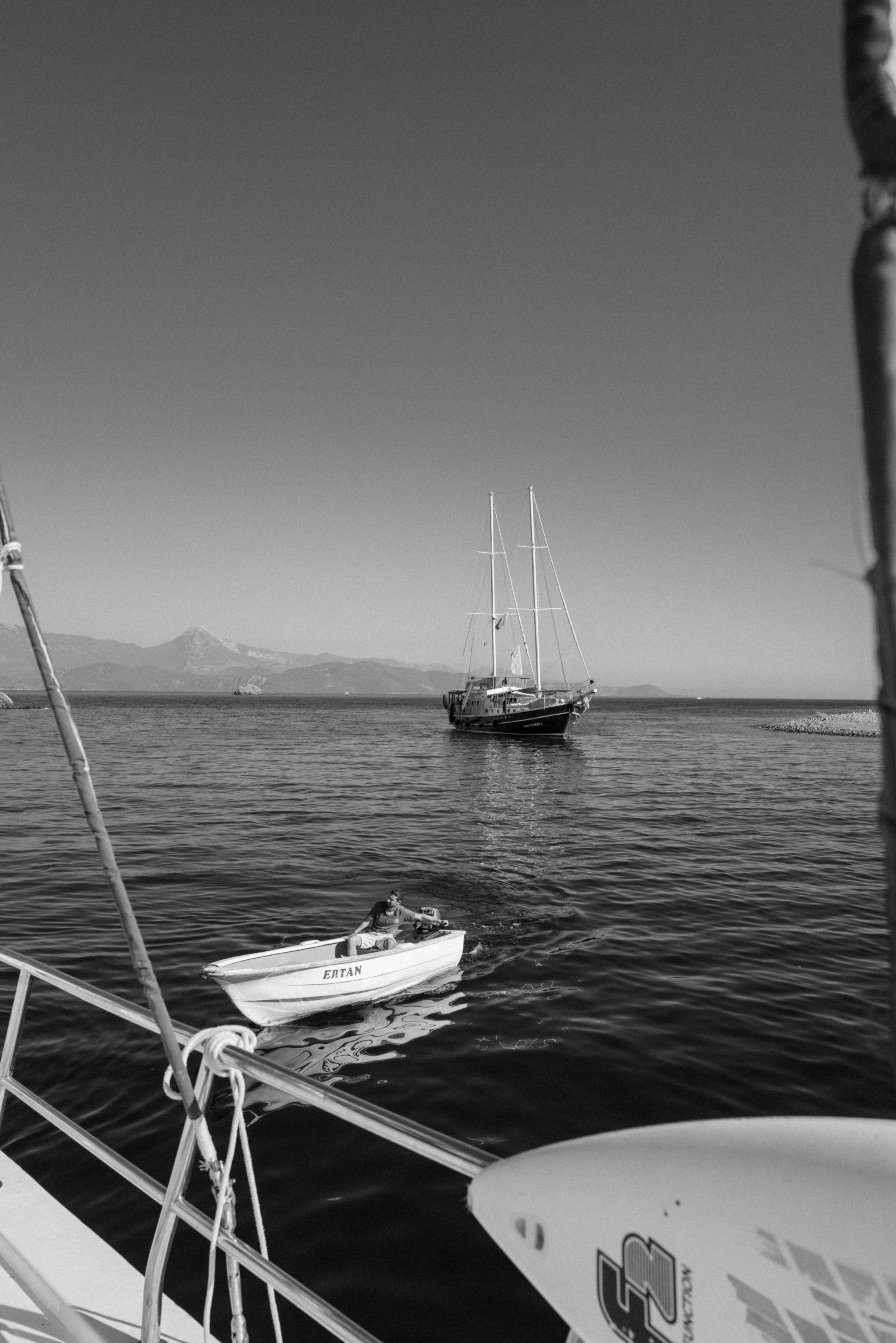 A monochrome sailboat rests beyond a dinghy on a calm mountain-framed bay.