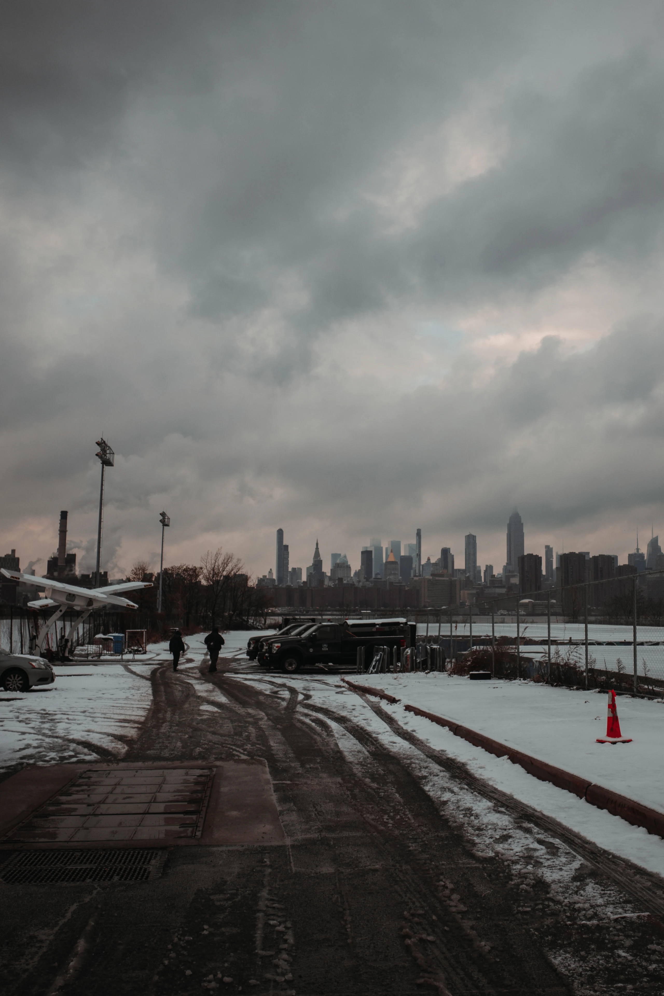 A snowy roadway and parked vehicles beneath dark clouds with the Manhattan skyline in the distance.