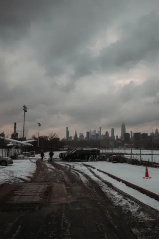 A snowy roadway and parked vehicles beneath dark clouds with the Manhattan skyline in the distance.