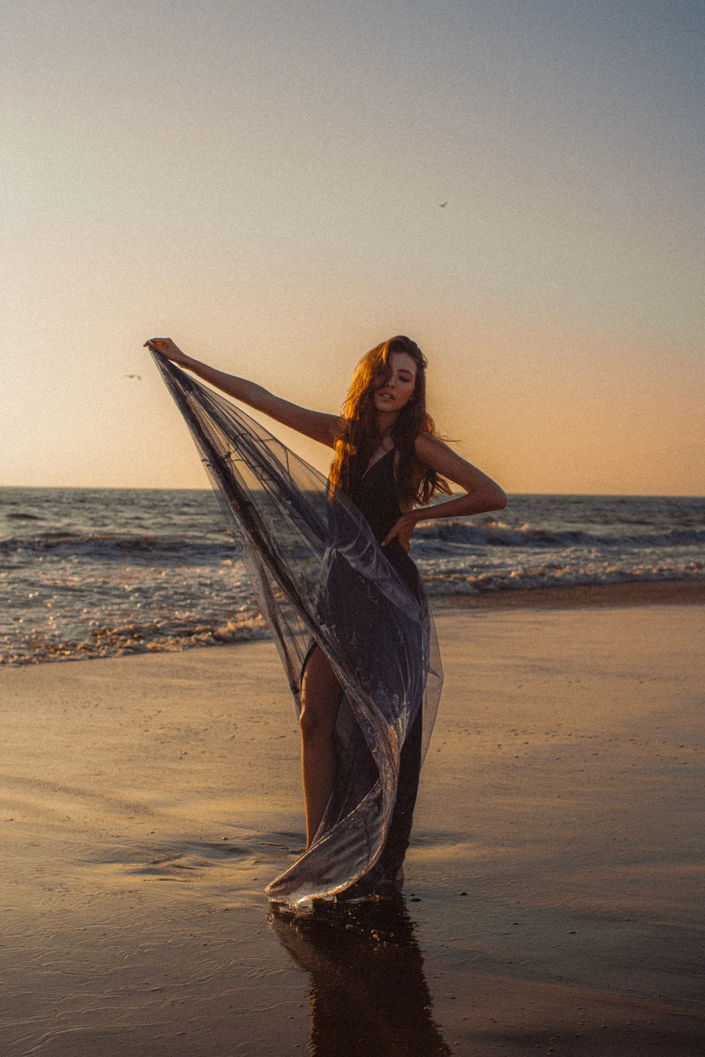 Woman holding flowing fabric along the shoreline at sunset.