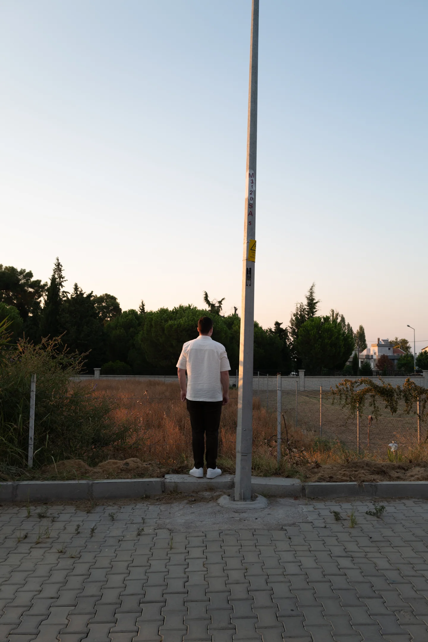 A man in a white shirt stands beside a tall streetlight at the edge of a cobbled street.