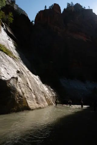 Hikers wade through the Virgin River beneath towering Zion canyon walls, with sunlight reflecting off wet rock on the left.