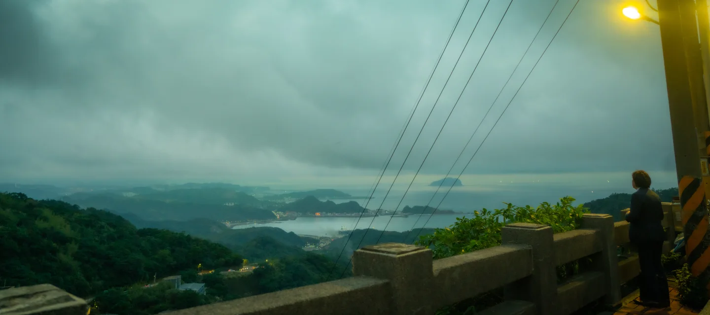 A person stands at a concrete lookout above a mountainous coast beneath low clouds.