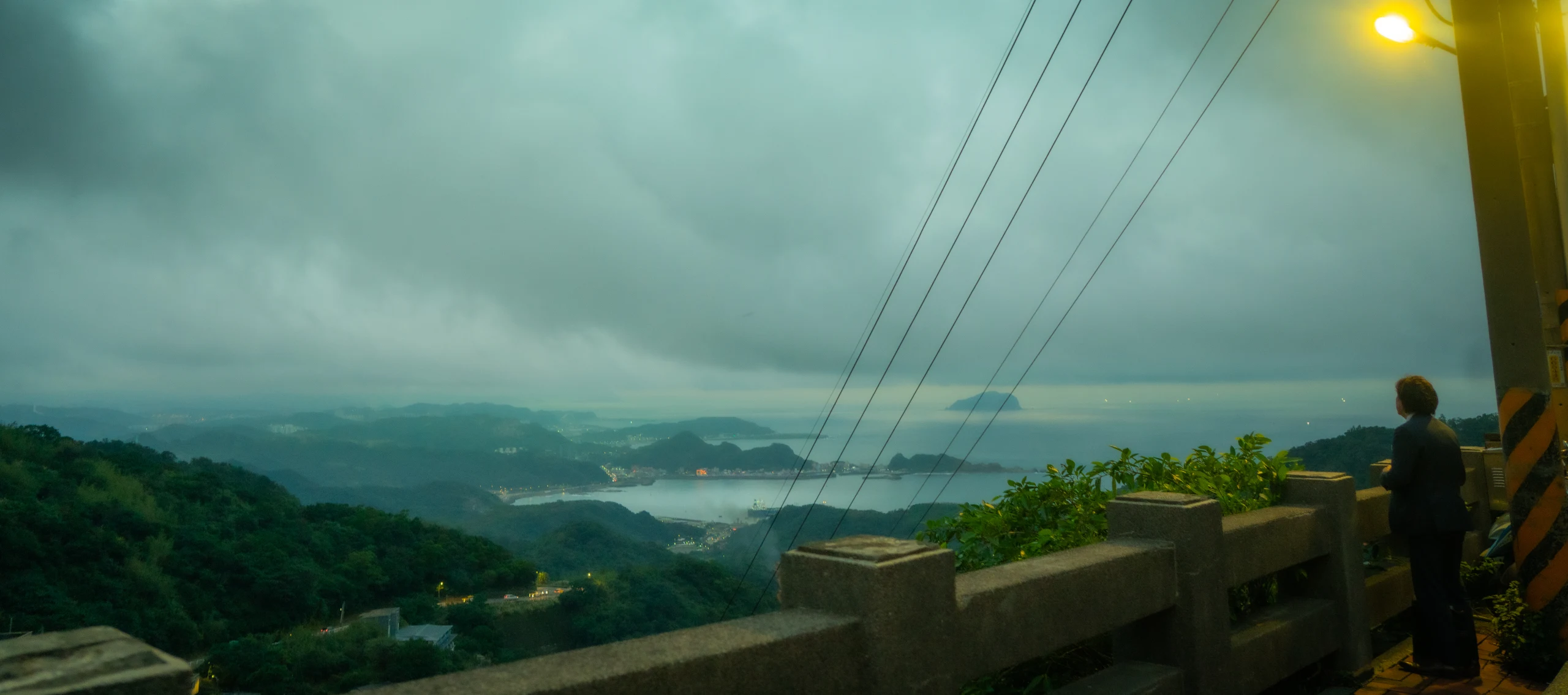 A person stands at a concrete lookout above a mountainous coast beneath low clouds.