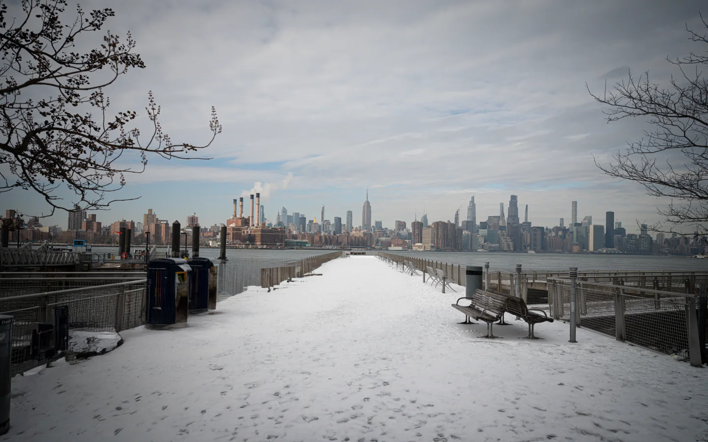 Snow-covered Williamsburg waterfront promenade with empty benches and the Manhattan skyline across the East River beneath gray clouds.