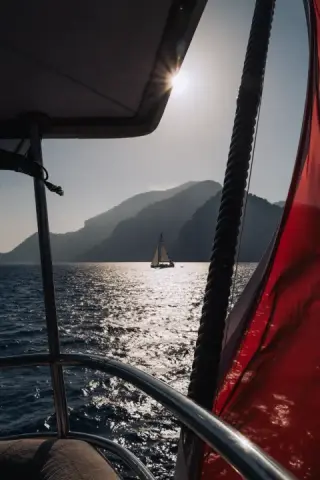 A distant sailboat crosses sparkling water framed by dark rigging and a red canvas panel.