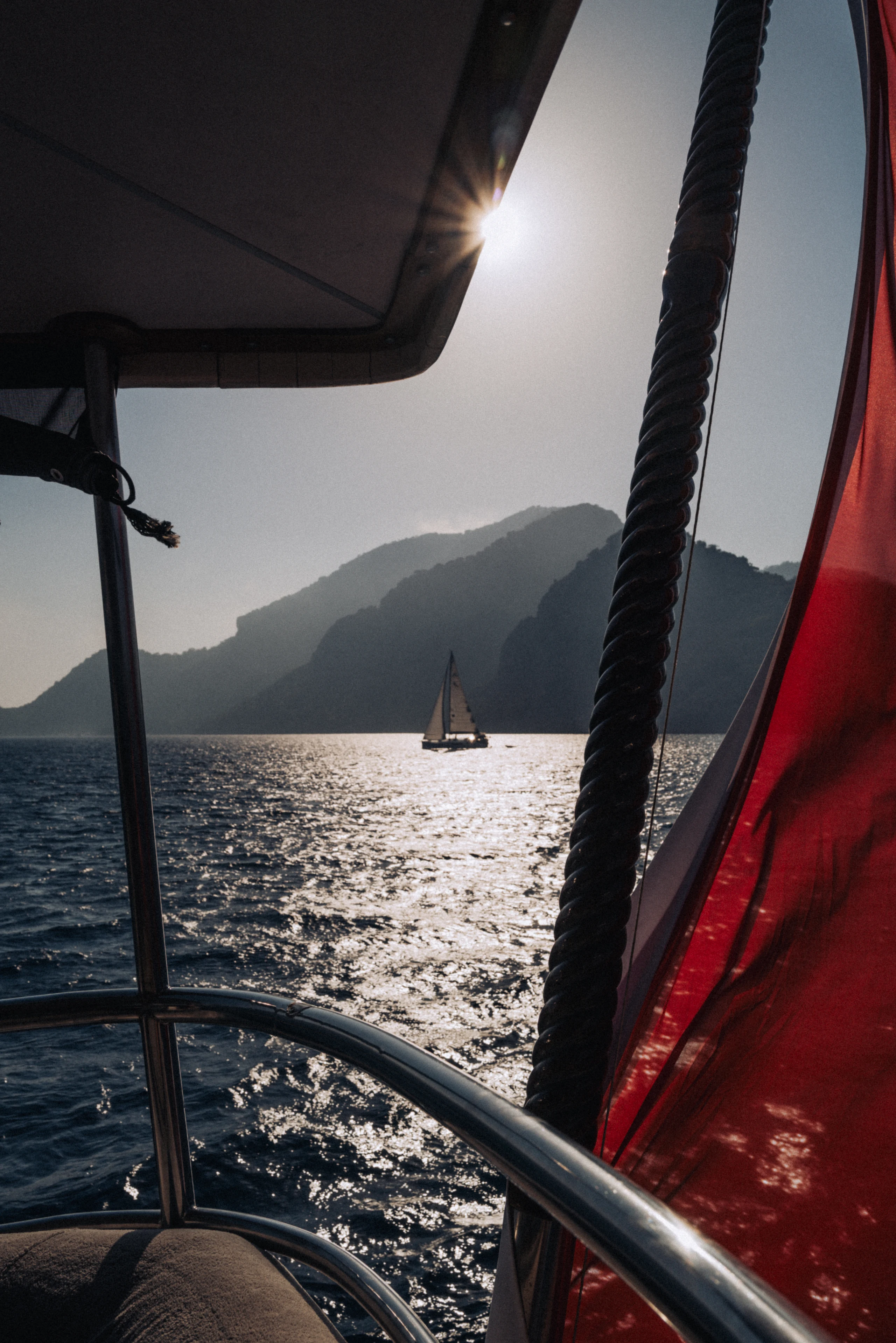 A distant sailboat crosses sparkling water framed by dark rigging and a red canvas panel.