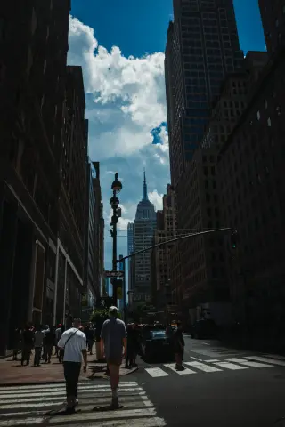 Pedestrians cross a shadowed Midtown street with the Empire State Building centered between tall buildings under blue sky and clouds.